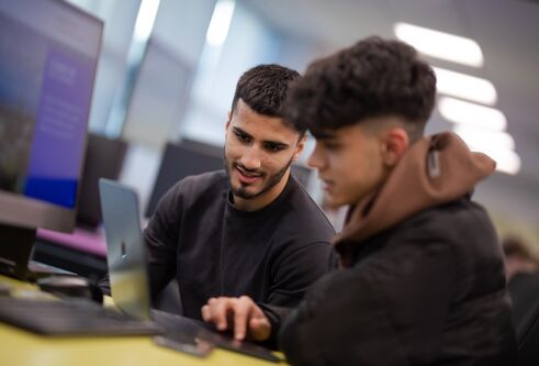 two students work on a laptop together, other computers are in the background along with large monitors