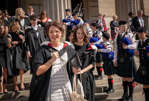 Two girls walking down steps outside of a building, wearing graduation robes. A group of graduates stand behind them, and a row of bagpipers stand lined up on the steps.