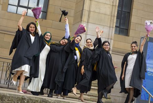 Seven Afghan women stand on the steps of a building wearing graduation robes and celebrating.