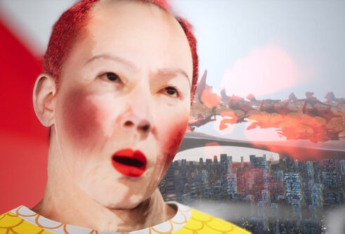 Digital video still. A close-up CGI rendering of an asian man with white make-up on his face, rouge on his cheeks and dyed short red hair. Over his should we can see two types of landscape. Rows of skyscrappers and above this red amorphous shapes floating in a digital pink hued sky.